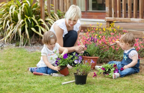 A woman with short blonde hair is sitting on a well-maintained lawn in a garden, engaging with two young children, both of whom are girls. The children are seated on the grass, each tending to small flower pots filled with vibrant purple, pink, and yellow flowers. The garden features a lush green grass area, bordered by a variety of plants with long, spiky leaves and leafy shrubs. In the background, there is a wooden deck with a railing, and the garden appears to be part of a residential property in Dulwich, South London, as suggested by the context of the webpage. The setting is outdoors on a bright, slightly overcast day, with natural light illuminating the scene. The overall environment showcases a tidy, landscaped garden space suitable for family activities and outdoor gardening, reflecting professional gardening maintenance and care provided by Garden Maintenance Dulwich for local homes.