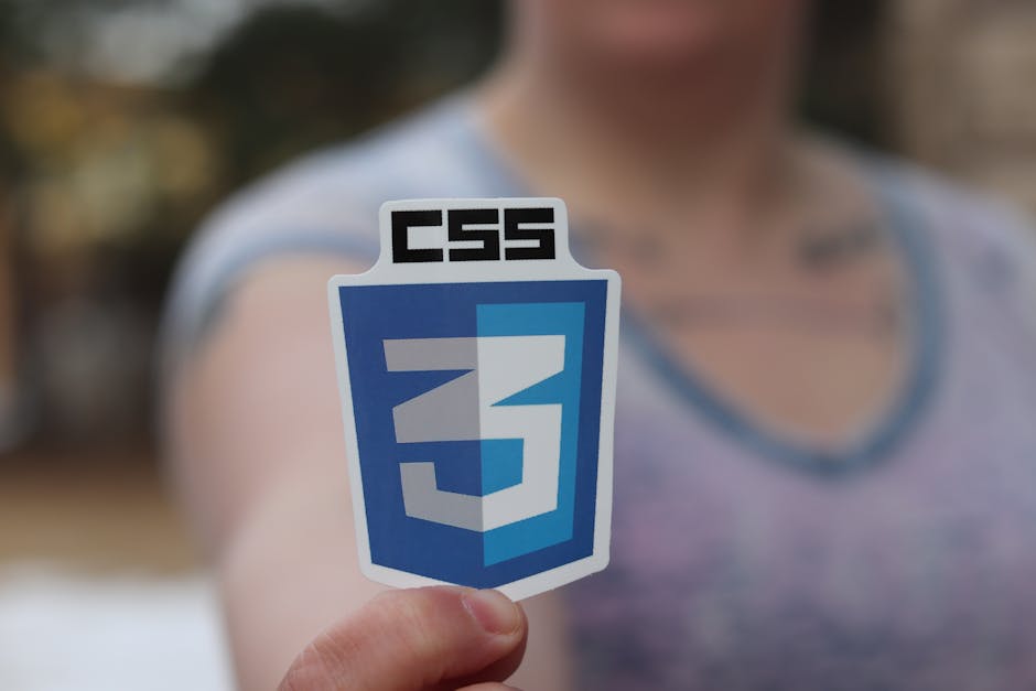 A person holding a CSS3 logo sticker is visible in the foreground, with the individual wearing a light grey T-shirt featuring a blue trim around the neckline. The background shows a well-maintained fr
